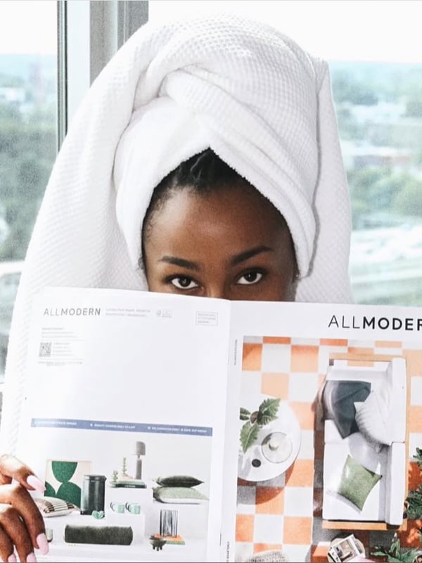Woman with towel wrapped around her head posing playfully in front of a window with city view, holding a colorful magazine.