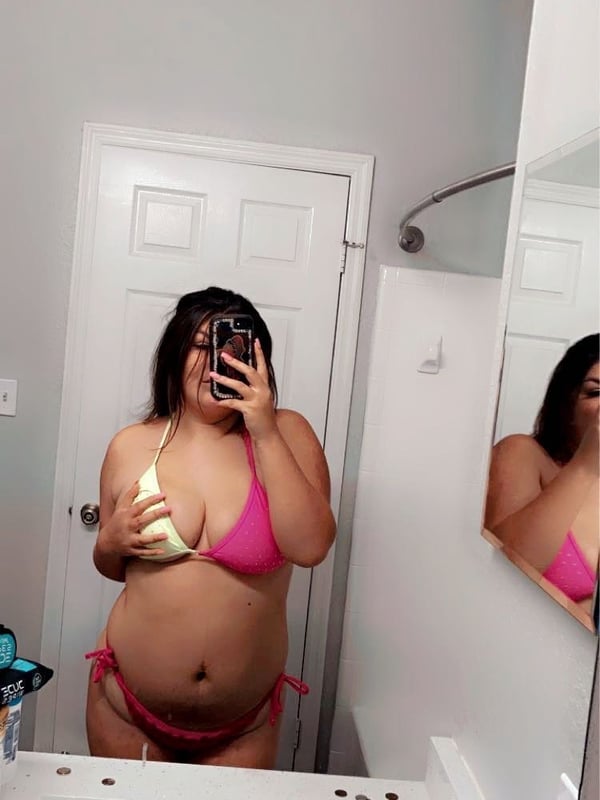 A woman taking a mirror selfie in a bathroom, wearing a pink bikini and holding a phone.