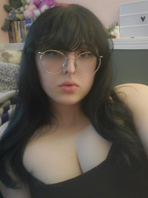 Young woman with long dark hair and glasses smiling at the camera in a cozy bedroom setting with plants and stuffed animals.