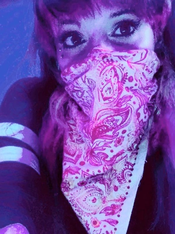 A young woman with purple hair wearing a pink patterned bandana over her face, posing in a vibrant neon-lit setting with colorful lights.