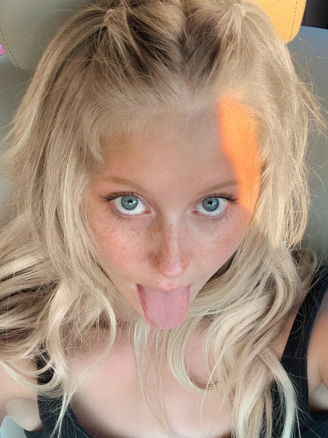 A young woman with blonde hair and freckles sticking out her tongue in a car selfie, seated with a pink drink nearby.