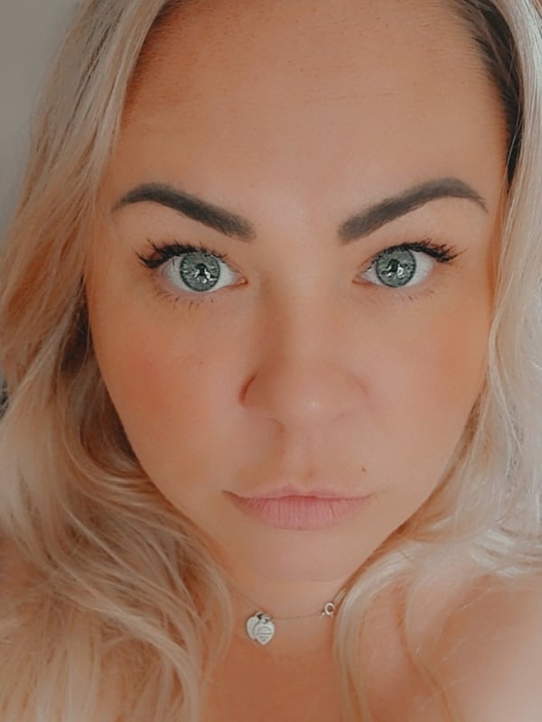 A close-up selfie of a woman with long blonde hair, green eyes, and a necklace, smiling at the camera.