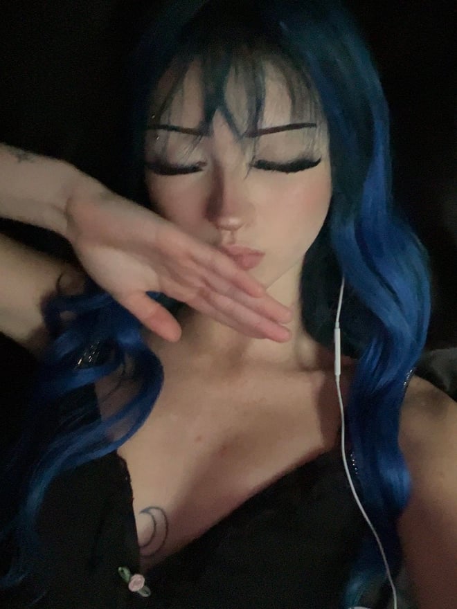 A young woman with long blue hair posing for a mirror selfie in a checked top.