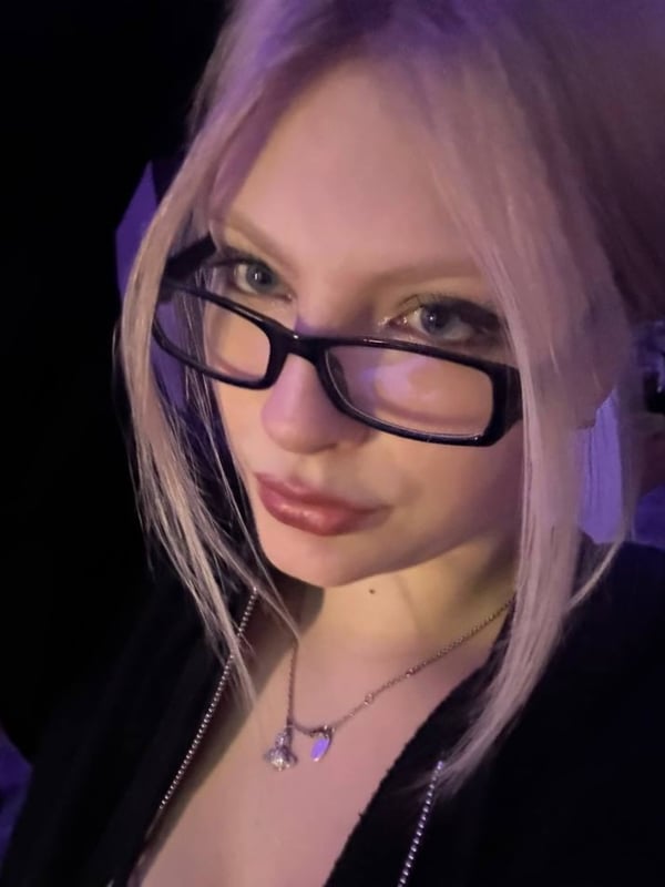 A young woman with blonde hair wearing black-framed glasses and a necklace, posing with a subtle smile against a dark background.