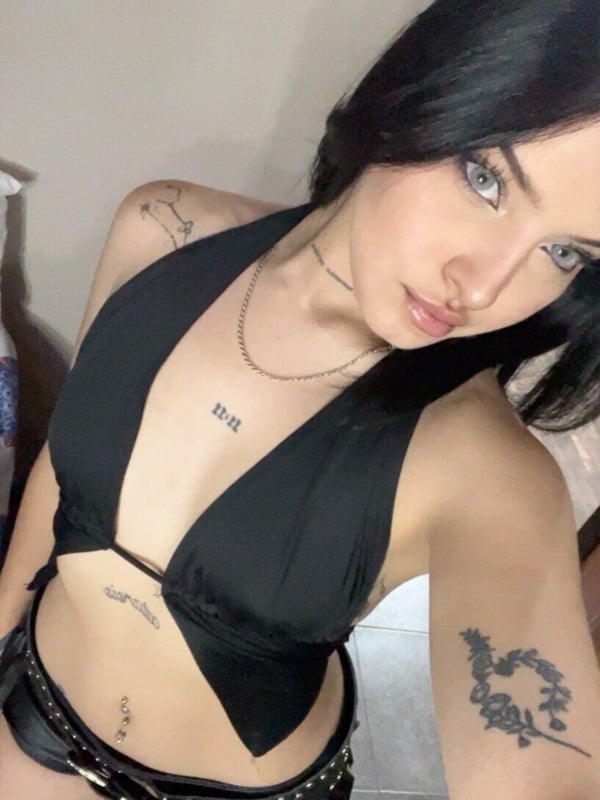Young woman with dark hair taking a mirror selfie in a black cropped hoodie and low-rise pants, standing in a tiled room with graffiti.