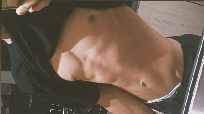 A young man posing shirtless in front of a mirror, lifting his shirt to show his toned torso.