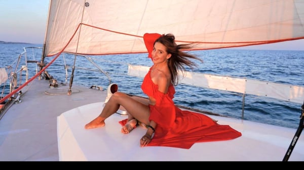 Woman in a flowing red dress posing elegantly on a yacht deck with sails and ocean backdrop at sunset.