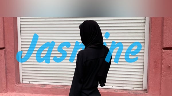 A person in modest black attire standing in front of a pink wall with a white shutter and blue text.