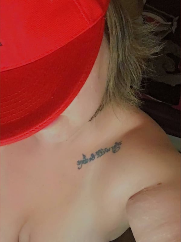 Close-up selfie of a woman wearing a red cap, showing her shoulder and collarbone area with a small tattoo.