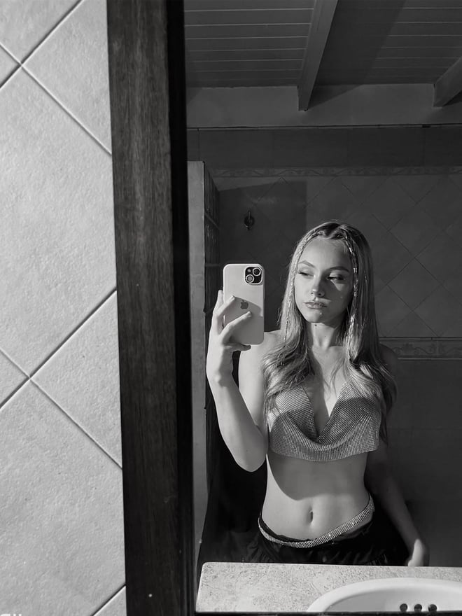 A young woman taking a selfie in a white satin crop top with a small tie detail, against a plain wall.