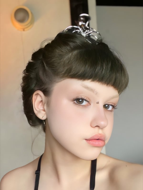 Close-up portrait of a young woman with dark bob hair styled with a shiny hair clip, wearing a top and subtle facial piercing, against an indoor background.