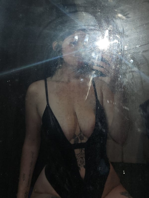A close-up selfie of a person in shiny black lingerie with tattoos visible, smiling in a reflective indoor setting.