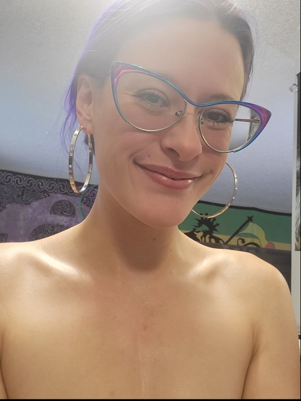 A young woman with purple hair and colorful glasses smiling at the camera, standing topless in a room with patterned walls.