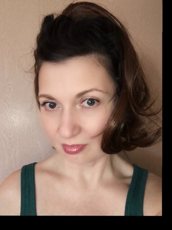 Woman with brunette wavy hair smiling confidently, wearing a green top against a neutral background.