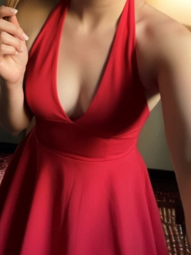 A woman smiling in a red halter dress, standing indoors with warm lighting from a lamp.