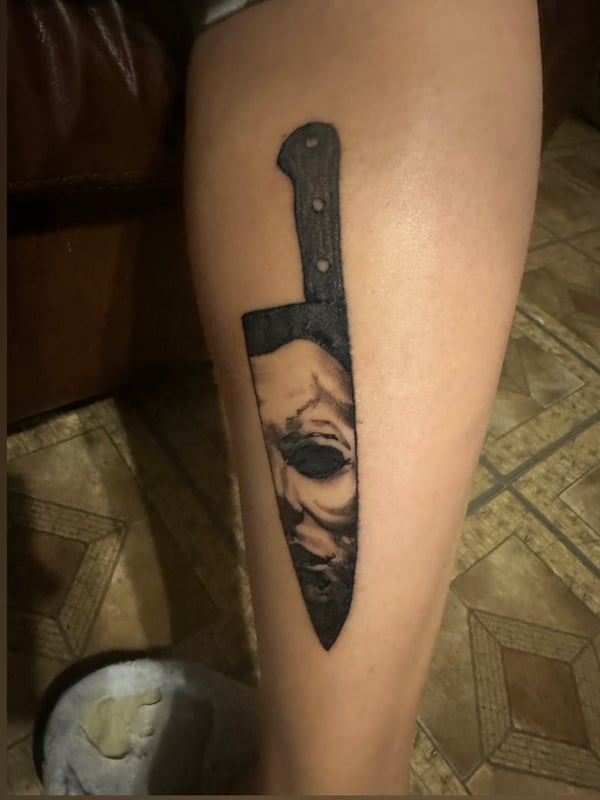 Close-up of a woman's leg displaying a detailed black tattoo of a knife with a masked face design.