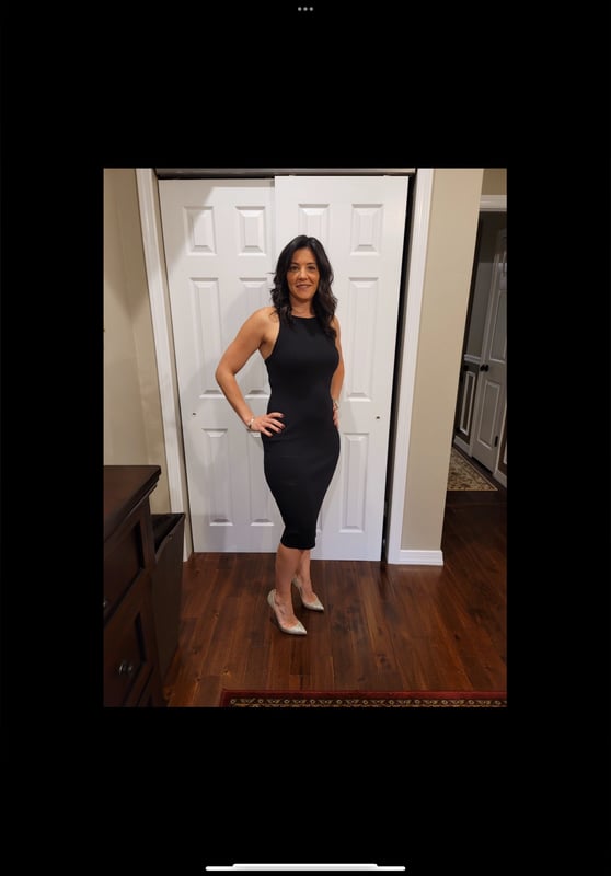 A woman in a sleek black dress posing confidently in front of a white door in a home interior with wooden flooring.