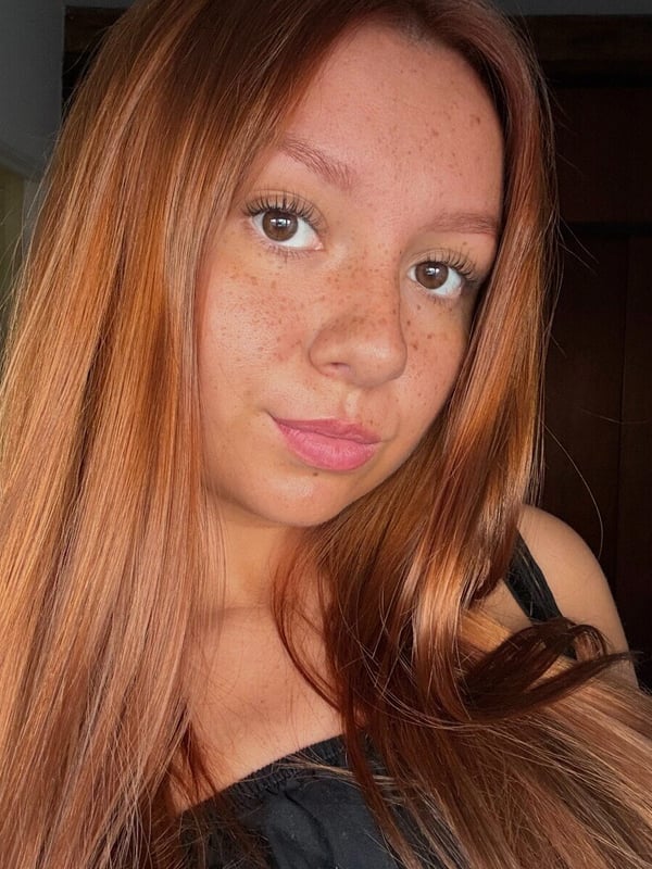Close-up selfie of a young woman with freckles and long red hair, smiling softly while wearing a black top, standing in a bedroom with wooden furniture.