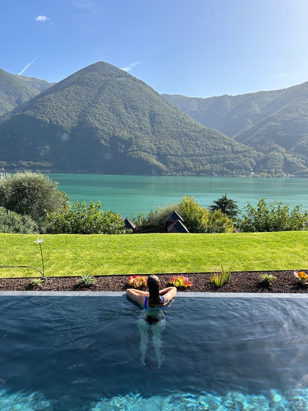A woman relaxing in a bikini at the edge of an infinity pool overlooking a turquoise lake and majestic mountains under a clear blue sky.
