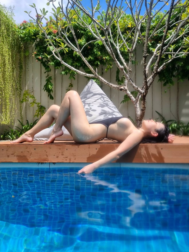 A woman lounging by a pool in a swimsuit, surrounded by tropical plants and a serene outdoor setting.