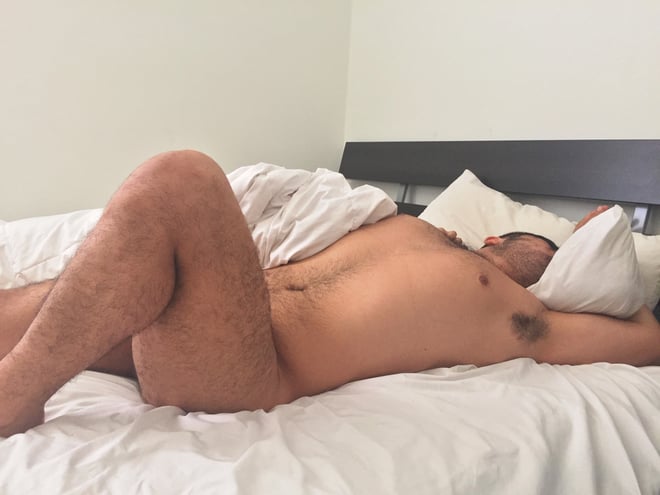 A man lying shirtless on a bed with white sheets and pillows in a bedroom.