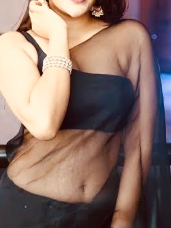 Woman smiling with hand on cheek, adorned with large earrings and pearl bracelet, dressed in a sheer black saree-style top against a dimly lit background.