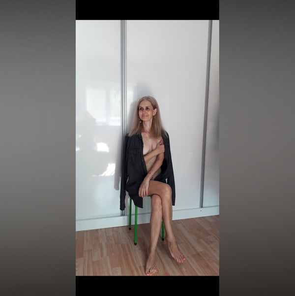 A woman with long blonde hair sits on a green chair in front of white wardrobe doors, wearing a dark blazer and posing with her arms crossed over her chest.