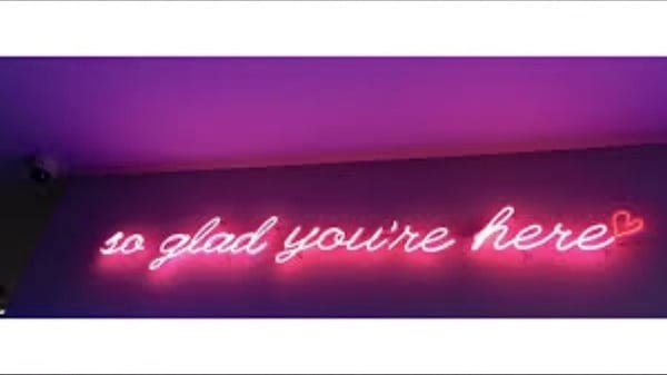 Pink neon sign glowing with the text 'so glad you're here' against a purple-lit background.