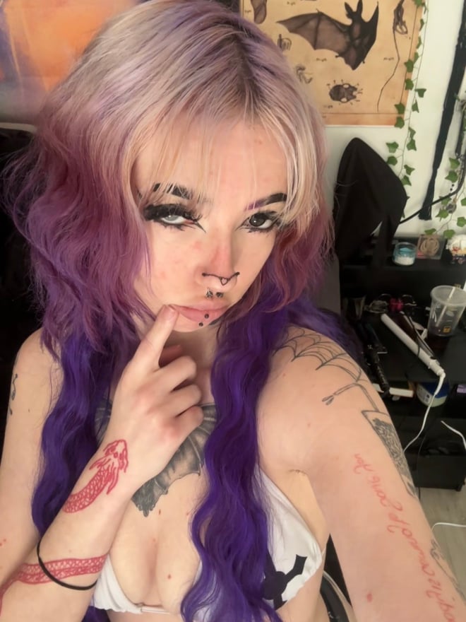 A close-up selfie of a person with purple hair and face tattoos looking at the camera.