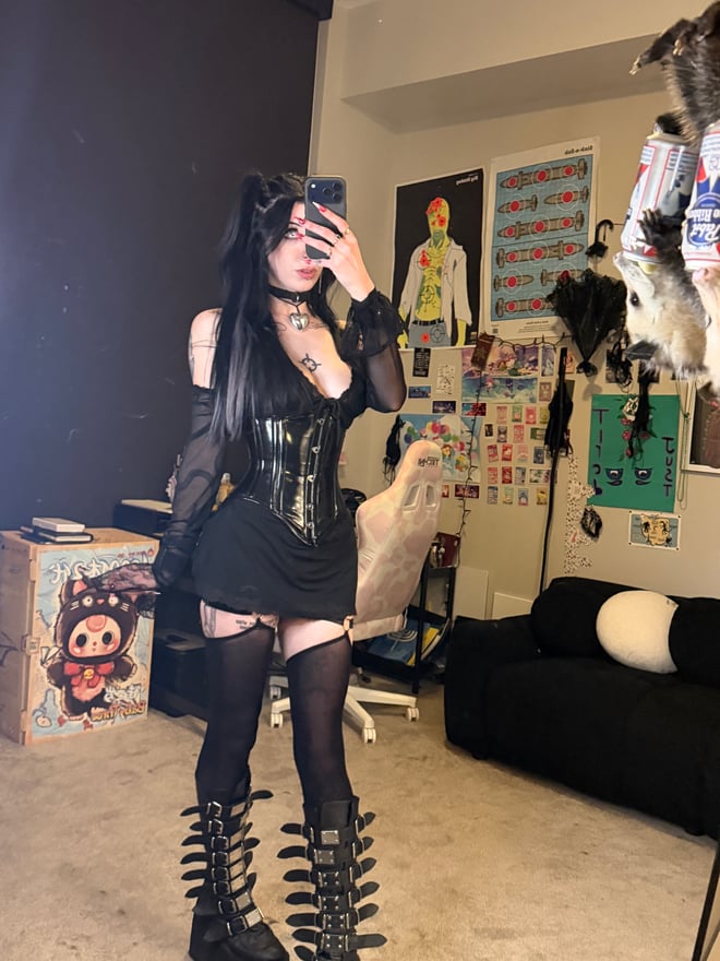 A person in alternative attire with cat ears and glasses posing playfully on a bed with pillows and soft lighting.