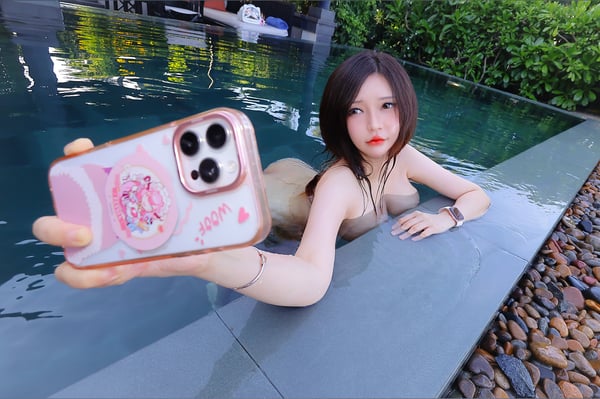 Young woman with dark hair sitting by a pool, holding up a pink phone case with anime designs while smiling at the camera.