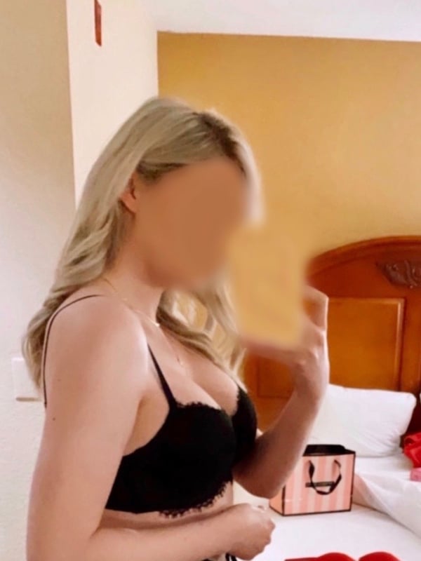 A blonde woman in a black bra standing in a bedroom with blurred face.