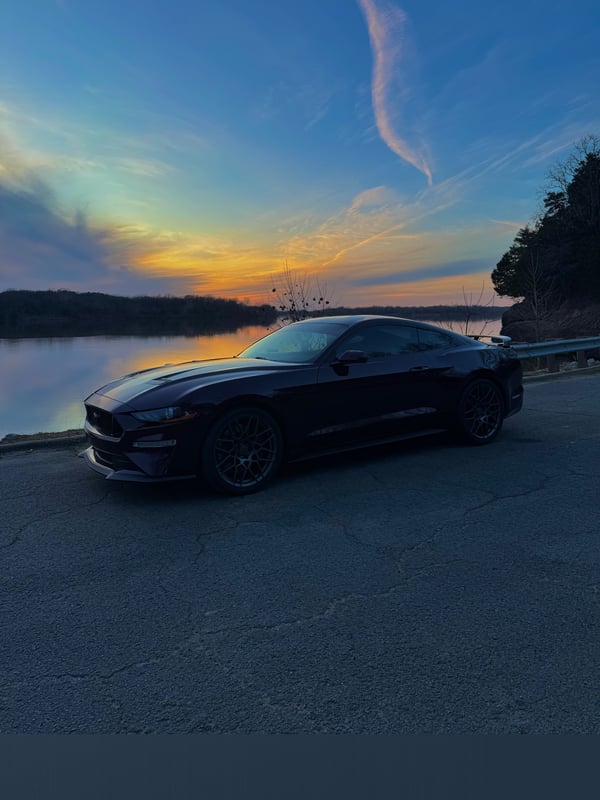 A sleek black sports car parked by a scenic lakeside at sunset with vibrant sky colors and surrounding trees.