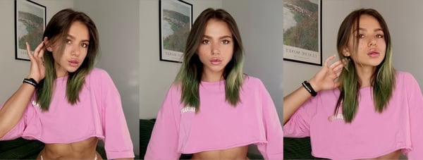 Young woman with green-tinted hair posing playfully in a pink cropped top inside a room with artwork on the walls.