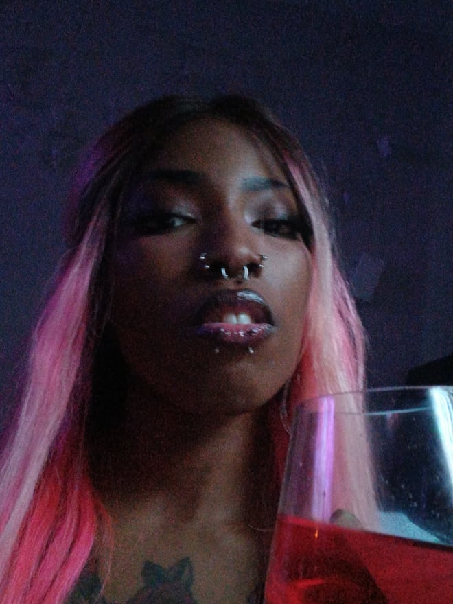 Close-up portrait of a woman with long pink hair and facial piercings, holding a glass of red drink against a dark background.