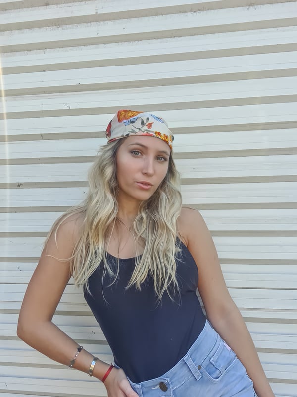 Young woman with long blonde hair wearing a colorful headscarf, black tank top, and light jeans, posing confidently against a white shuttered wall.