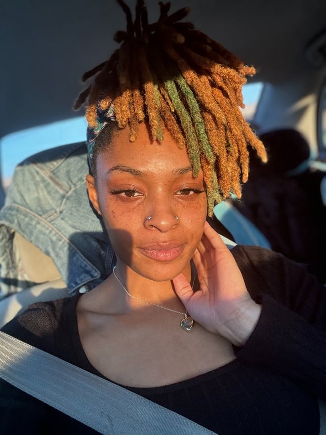 A young woman with colorful dreadlocks sits in a car, touching her face while wearing a black top, with natural light illuminating her features.