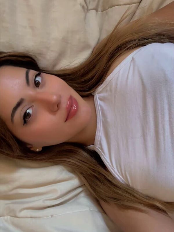 Young woman with long blonde hair lying on a bed, wearing a white tank top, smiling at the camera.
