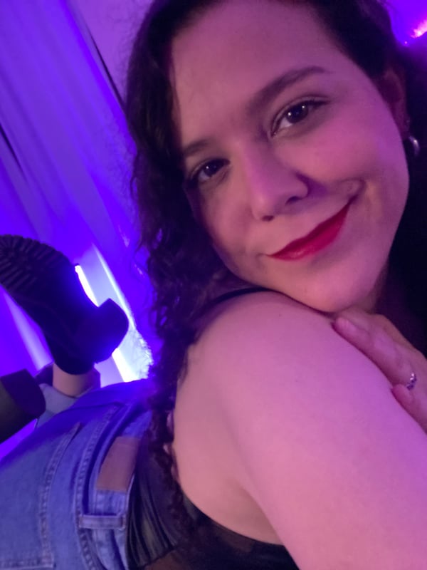 Young woman with curly dark hair and red lipstick smiling at the camera, reclining on a bed in a purple-lit room while wearing jeans and a tank top.