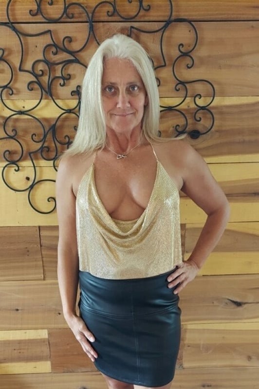 A mature woman with long blonde hair posing confidently in a gold chainmail top and black skirt against a wooden wall with decorative elements.
