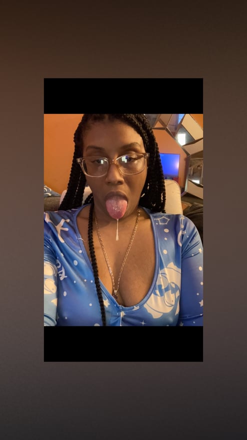 A woman with braided hair wearing glasses and a blue outfit, posing playfully indoors with warm lighting.