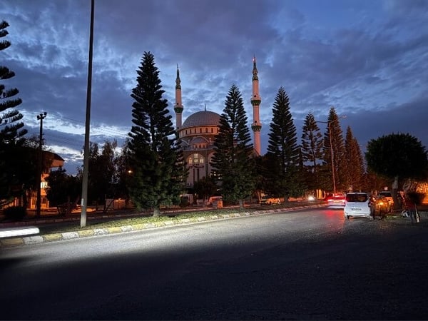 Scenic view of a mosque with twin minarets at dusk, surrounded by trees and streetlights along a road.