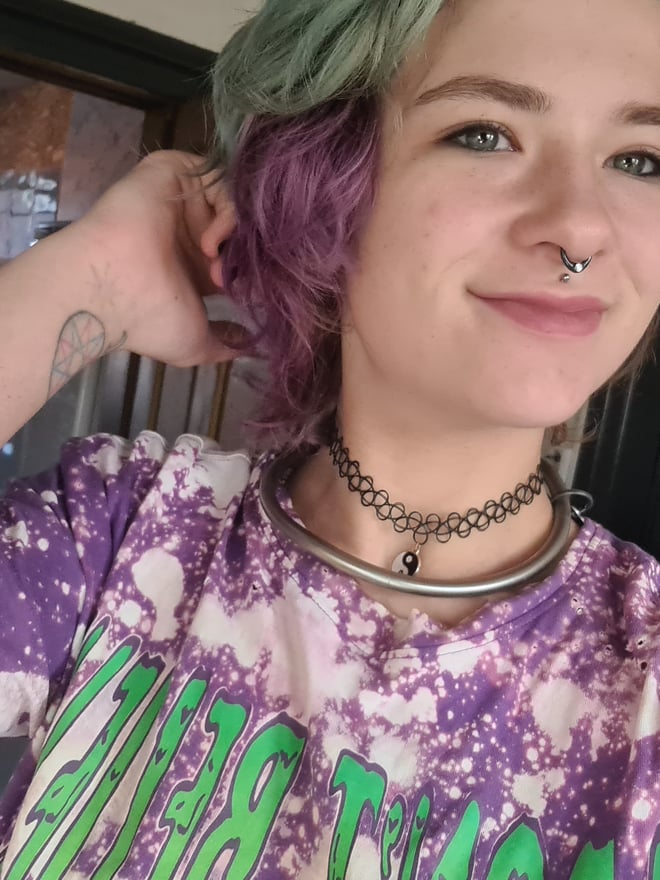 Young woman with dyed green and purple hair, nose piercing, and tattoos, smiling while wearing a purple tie-dye shirt and choker necklace indoors.