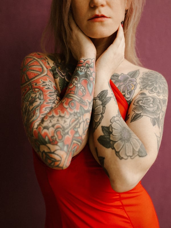 A woman with blonde hair and extensive tattoos on her arms stands against a purple background, wearing a red dress and touching her neck.