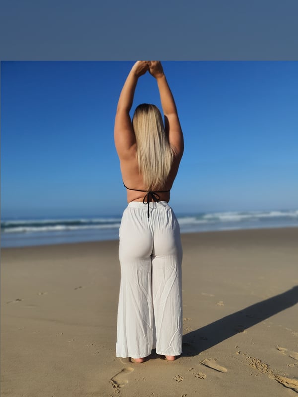 A woman with long blonde hair stands on a sandy beach, stretching her arms overhead while wearing a white bikini top and flowing white pants against a blue sky and ocean backdrop.