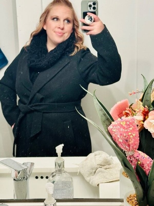 A smiling woman with blonde wavy hair taking a selfie in front of a mirror, dressed in a black coat and scarf, with decorative plants nearby.