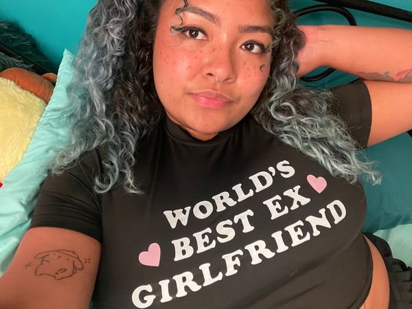 Young woman with colorful hair lying on a bed, wearing a black t-shirt with pink heart graphics and text, posing with a playful expression.