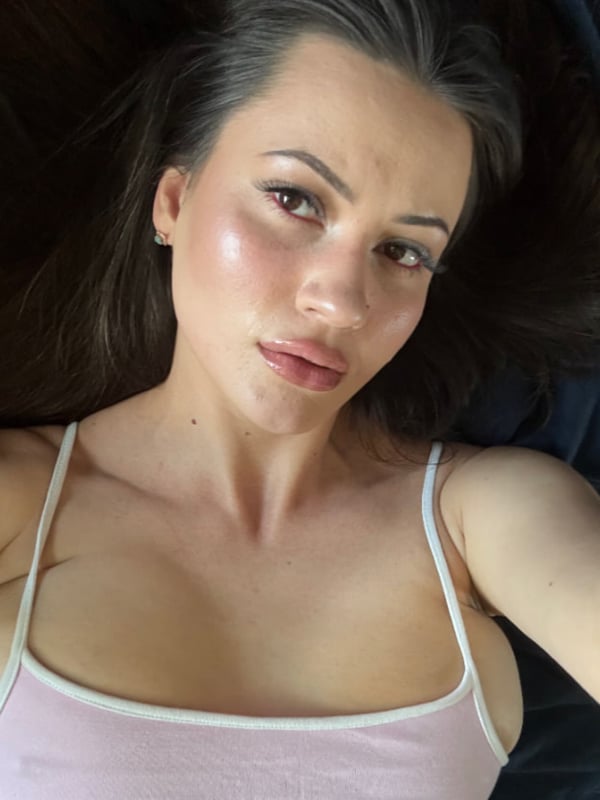 A woman lying on a dark surface taking a selfie, wearing a pink spaghetti strap top, with long dark hair and subtle makeup.