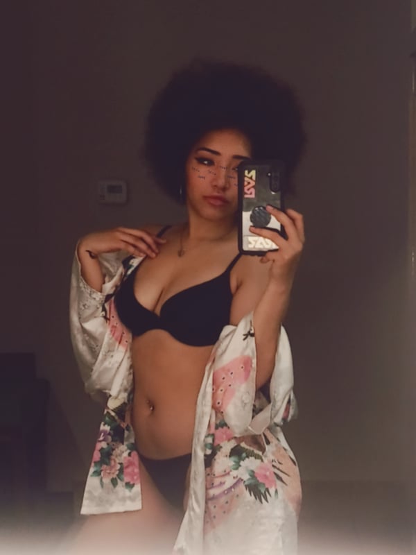 A woman with curly hair posing in a black bra and open floral robe, holding her phone for a selfie.