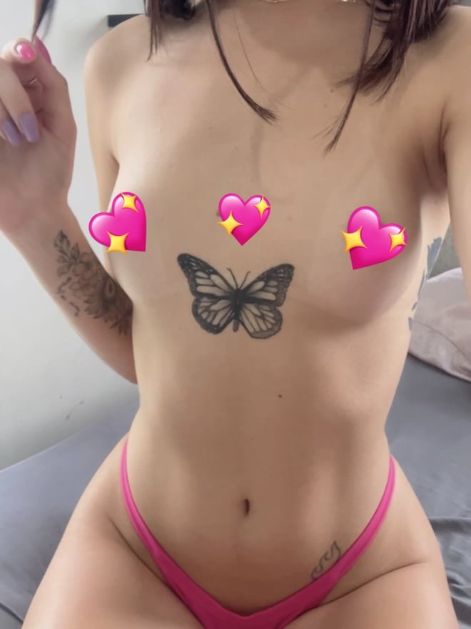 A young woman with dark hair posing indoors, displaying butterfly and other tattoos while wearing pink attire.